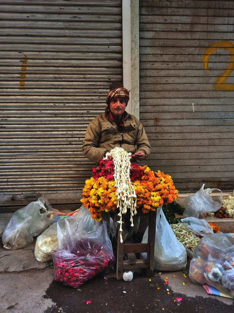 A flower vendor in a lane of Old Delhi, India. The vendor is selling a tapestry of flowers, including roses and marigolds. The flowers are arranged in a beautiful and colorful display, and the vendor is surrounded by locals who are browsing the flowers. This is a scene from an unexplored part of Old Delhi that is full of local color and authenticity. It is a great place to find unique and beautiful flowers that you won't find anywhere else. This is a popular spot for tourists on our food and photography walking tour in Delhi.